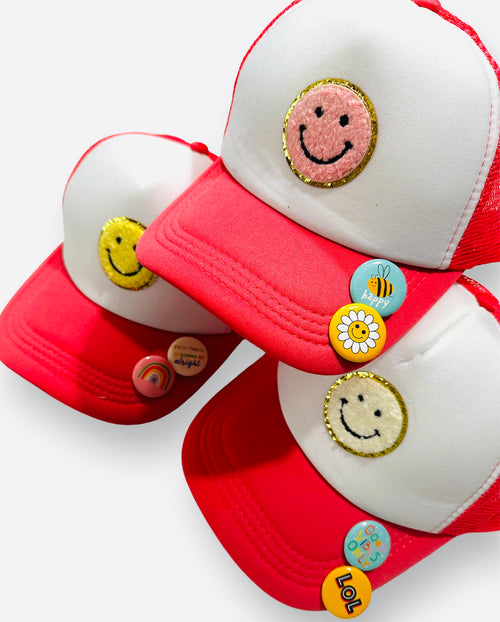 A white and bright red trucker hat with a smiley face patch and fun pins.