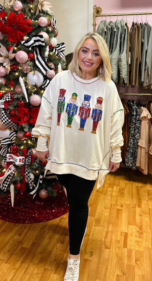 A white crewneck sweatshirt with multicolored stitching along with 4 sequin nutcrackers on the front.