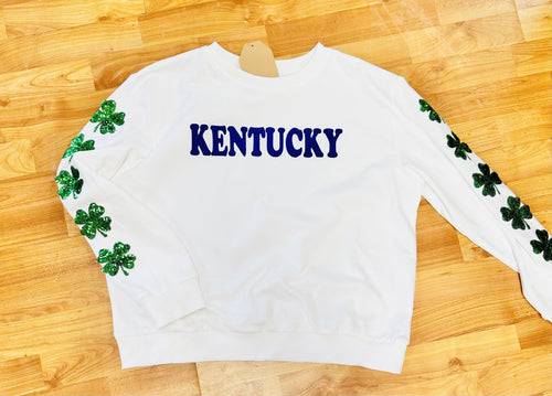 A white long sleeve crewneck with "Kentucky" written across the chest in royal blue along with green sequin clovers down each sleeve.