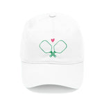 A white baseball cap with a green line drawing of pickleball paddles and a small pink heart above them.
