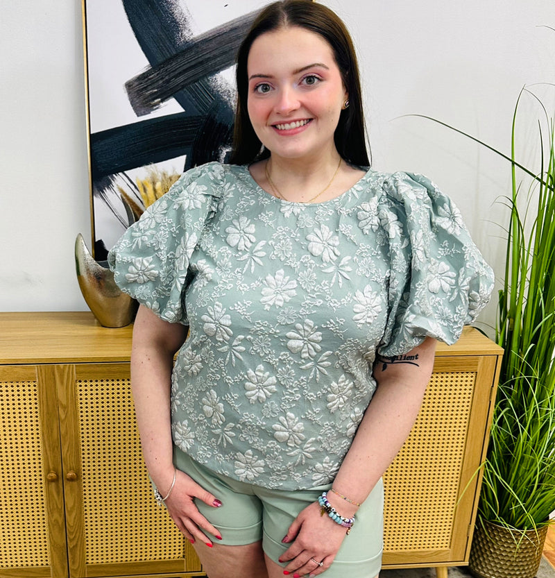 A muted green, short-sleeve blouse with a white floral print and puff sleeves.
