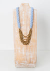A light blue and gold necklace featuring a light blue knit braid around the neck, then gold layered chains as the pendent. 