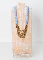 A light blue and gold necklace featuring a light blue knit braid around the neck, then gold layered chains as the pendent. 