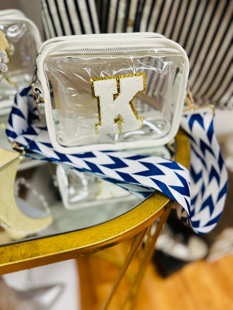 A small clear crossbody purse with a "K" patch in white and gold along with a blue and white chevron strap.