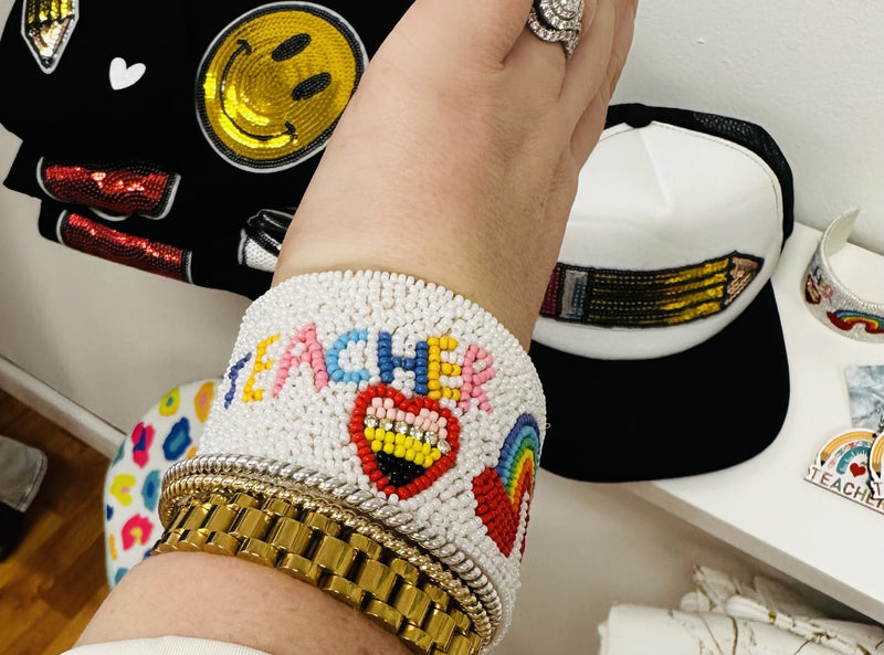 A white wide beaded bracelet cuff with "teacher" in multicolored letters and other school graphics.